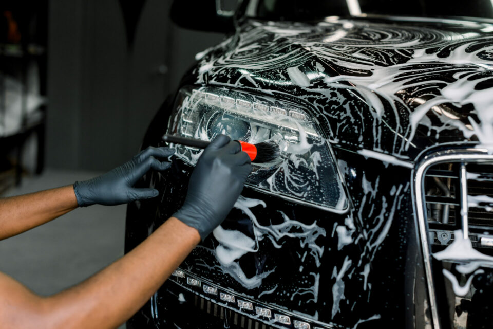 Close up photo of hands of auto service male worker in black protective gloves cleaning car hood and headlights with foam and soft brush. Carwash and detailing.